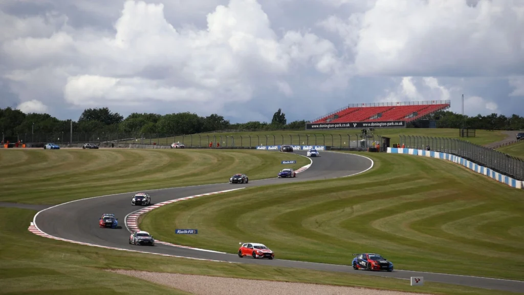 Touring cars race around a bend at Donington Park racetrack in the UK, with a red grandstand and dramatic skies in the background.