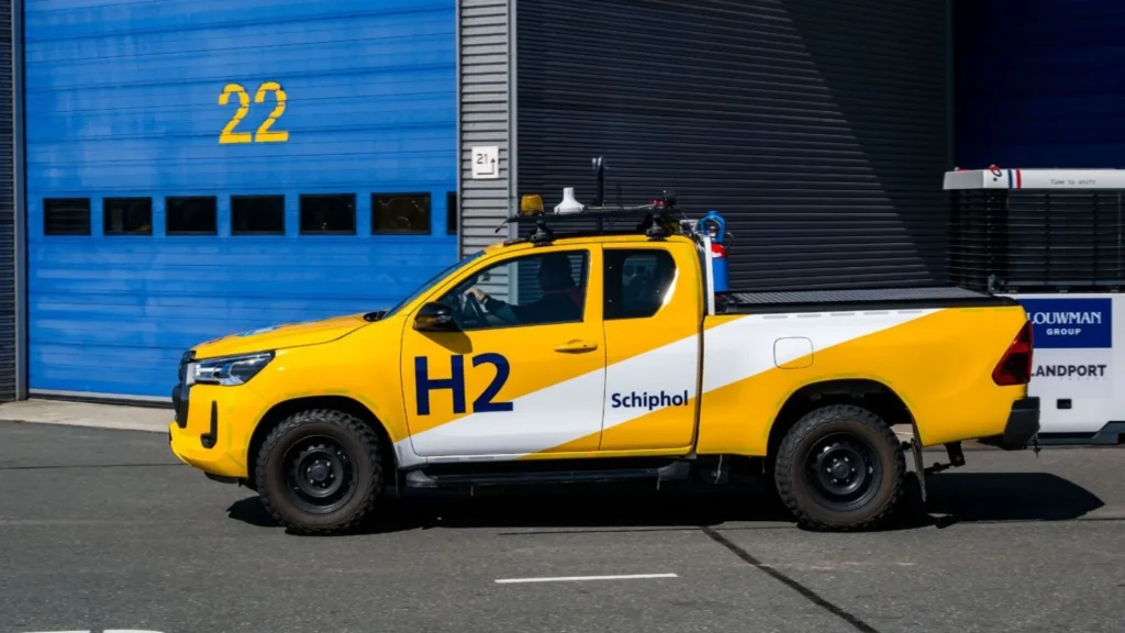 Yellow hydrogen-powered Toyota Hilux driving past maintenance buildings at Schiphol Airport.
