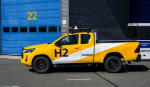 Yellow hydrogen-powered Toyota Hilux driving past maintenance buildings at Schiphol Airport.